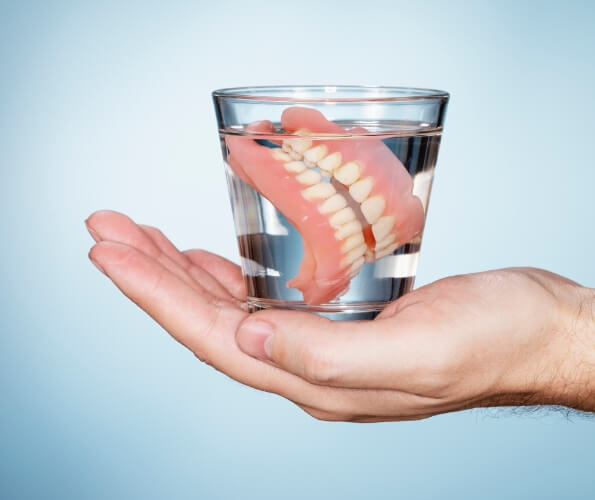 Hand holding a glass of water with dentures soaking inside of it