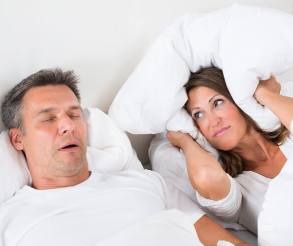 Woman covering her ears with pillow next to snoring man